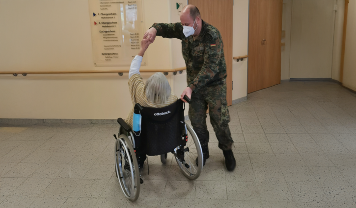 Man sieht einen Soldaten, der eine alte Dame im Rollstuhl durch das Seniorenheim begleitet