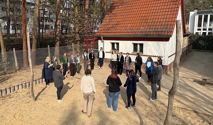 Auf dem Bild sind die Teilnehmenden des AWW-Regionaltages in einem Kreis stehend auf dem Gelände der Kita Wolkenflitzer in Berlin zu sehen.