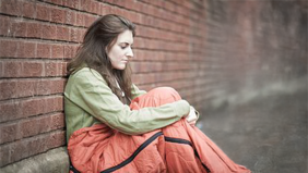 Das Bild zeigt eine obdachlose junge Frau vor einer Mauer sitzend