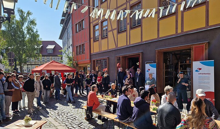 Das Bild zeigt Menschen, die draußen an einer langen Tafel sitzen und viele, die stehend einer Vorstellung zuschauen
