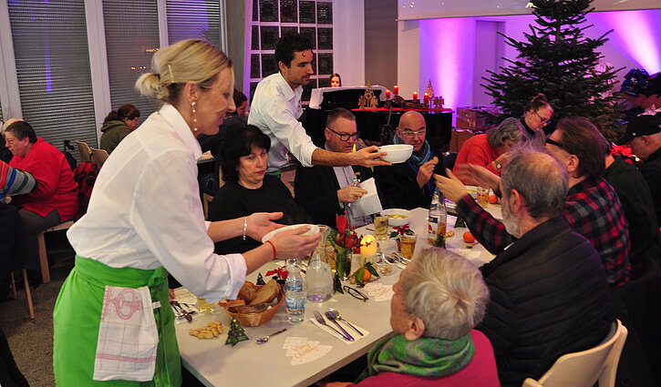 Gäste des Weihnachtsessens in der Adventgemeinde Ludwigsburg sitzen an festlich gedeckten Tischen und werden mit leckerem Essen bedient.